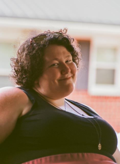 The owner of Azella Renae smiles in a vintage film photo. She has curly, short brown hair and green eyes. She is wearing a black tank top and layered necklaces.